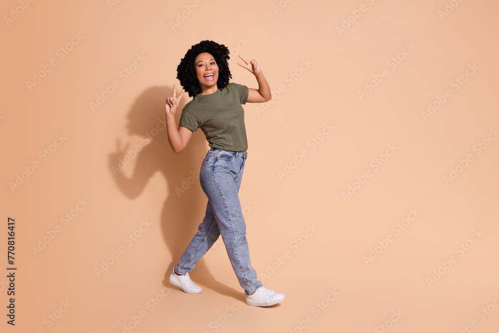 Full length photo of cheerful woman wear khaki t-shirt jeans trousers show v-sign go empty space isolated on beige color background Full length photo of cheerful woman wear khaki t-shirt jeans trousers show v-sign go empty space isolated on beige color background