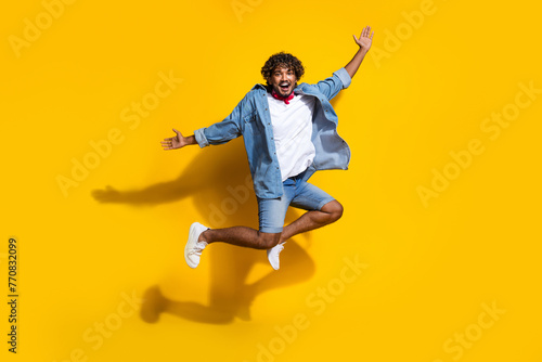 Full length photo of handsome young guy run jump spread hands dressed stylish jeans garment red scarf isolated on yellow color background
