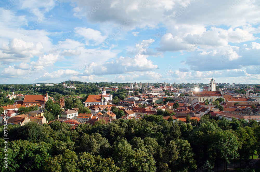 Top view from the mountain of the architecture of the old city of Vilnius with old buildings with orange tiled roofs, town halls, churches, towers, cathedrals, green deciduous forest, sunlight, clouds