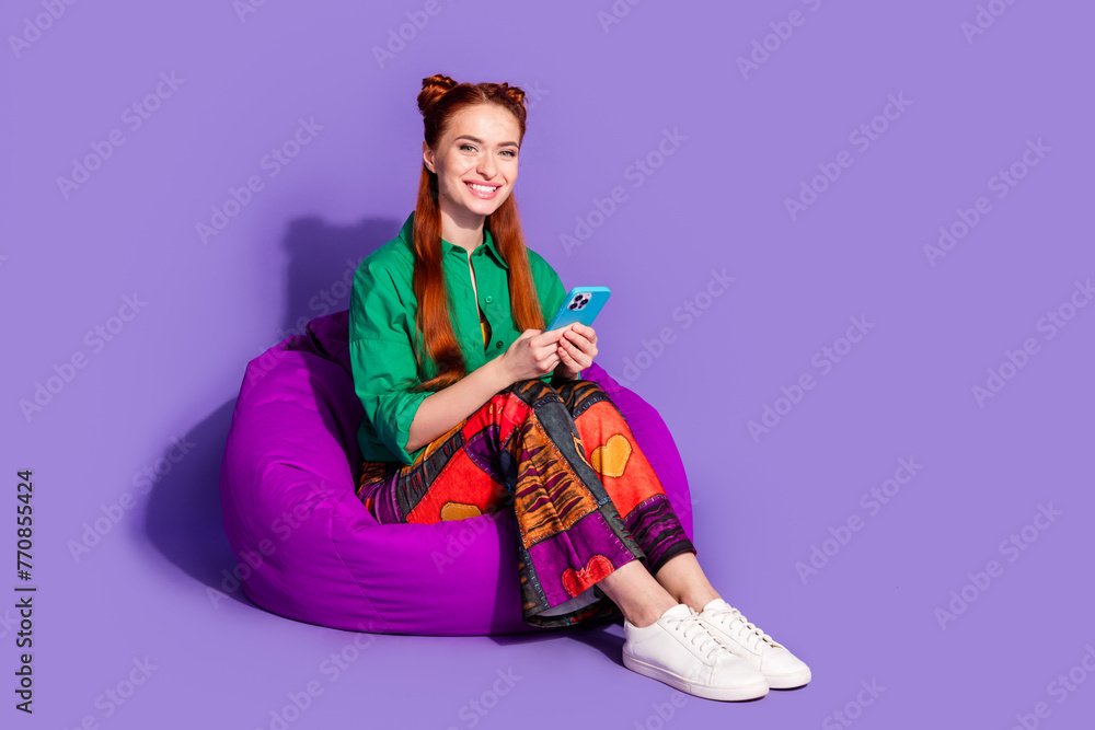 Full length photo of optimistic girl wear print vintage pants sit on pouf hold smartphone chatting isolated on violet color background Full length photo of optimistic girl wear print vintage pants sit on pouf hold smartphone chatting isolated on violet color background
