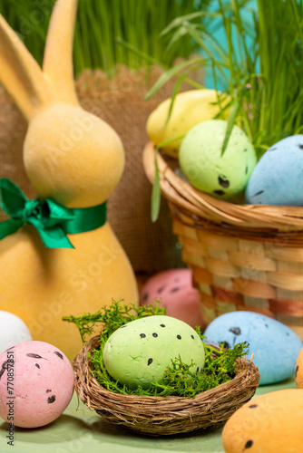 Easter decorative eggs in nest with moss, in basket, on table with yellow bunny and green grass.