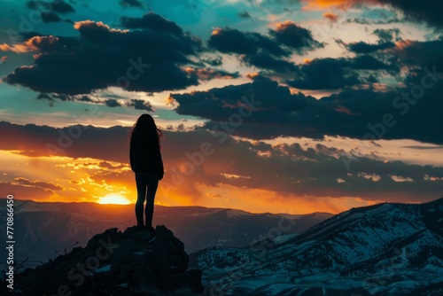 Silhouette of person against sunset sky - A lone person stands atop a mountain ridge, silhouetted against the vibrant hues of a sunset sky, overlooking a snowy landscape
