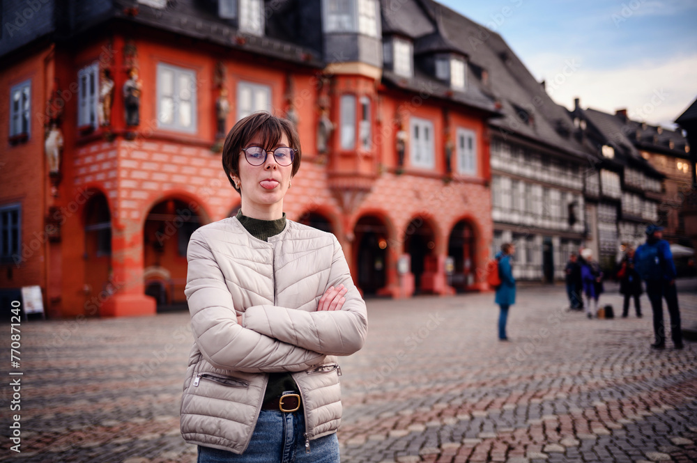 Fototapeta premium A woman stands confidently in a historic town square, her arms crossed, with traditional architecture creating a charming backdrop.