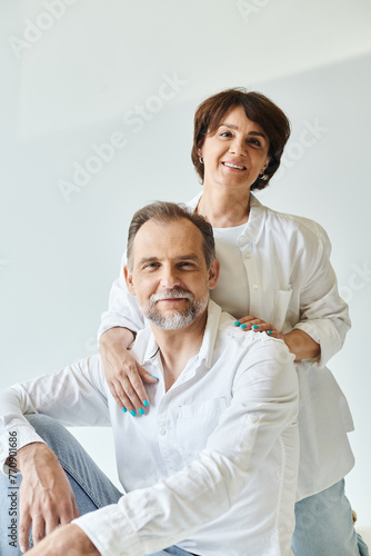 Middle aged smiling woman hugging her husband on grey background and looking at camera