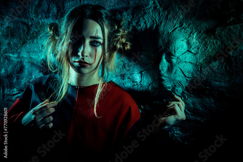 Halloween horrors. A scary clown girl, dressed in a red circus dress, in front of scary wall with stone faces.