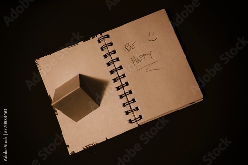 notepad with pages recycled from natural products, with the writing "be happy" and a smiling face, on the side a small pyramid made of the same material as the pages, which cast a shadow like an arrow