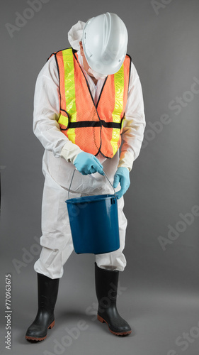 an unidentified person likley a man in a hazmat suit with rubber gloves and rubber boots with a hard hat and safety vest holding a pail isolated on grey photo