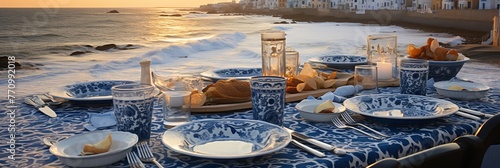 Beach dinner setup with bluewhite tablecloth, overlooking ocean. Table set for four, complete with plates, glasses, and cutlery. Wine, bread, and golden sunset complete the scene. photo