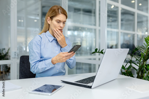 Fotografie Concerned professional female in a blue shirt at her office desk looking perplexed at her smartphone with a laptop and tablet nearby