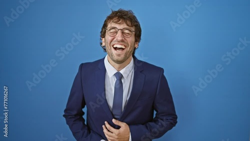 Joyful young man in a business suit laughs out loud, flashing a bright, handsome smile and clasping his hands in sheer delight! isolated against blue background.