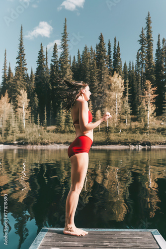 Woman in red swimwear running on a dock by a calm lake with a forest backdrop.