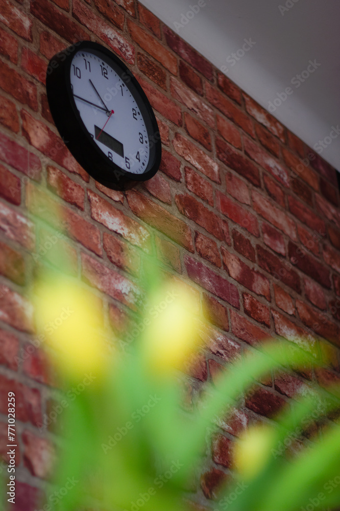 zegar okrągły biało czarny na ceglanej ścianie, z przodu rozmyty bukiet kwiatów żółto zielonych - obrazy, fototapety, plakaty 