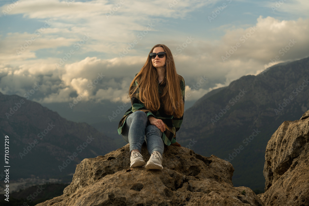 A woman with long hair is sitting on a rock in the mountains