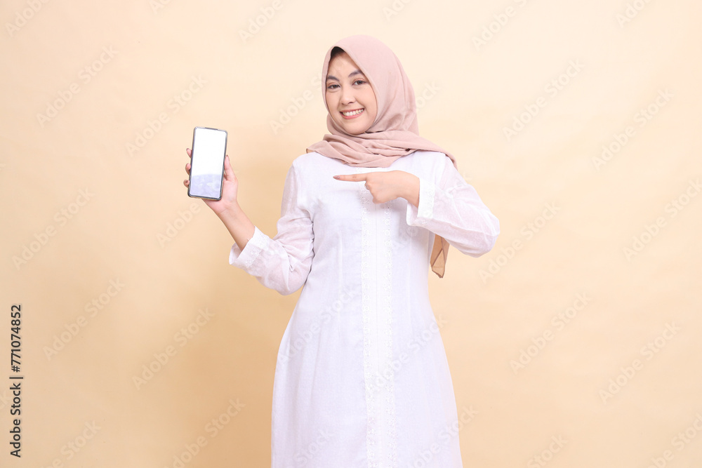 Young woman indonesia muslim wearing a hijab smiles cheerfully holding a smartphone gadget to the right in her hand while pointing at it. Lifestyle, business and commercial concept Young woman indonesia muslim wearing a hijab smiles cheerfully holding a smartphone gadget to the right in her hand while pointing at it. Lifestyle, business and commercial concept