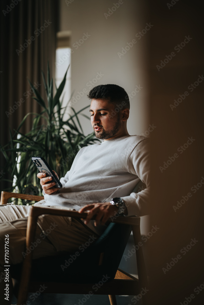 A young adult male relaxes in a chair, immersed in his smart phone in a cozy home setting.
