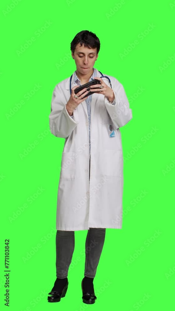Front view General practitioner playing video games on mobile phone app, having fun with online gaming competition against greenscreen backdrop. Cheerful doctor relaxing with internet game. Camera A. Front view General practitioner playing video games on mobile phone app, having fun with online gaming competition against greenscreen backdrop. Cheerful doctor relaxing with internet game. Camera A.