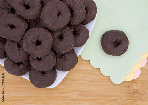 Cropped angle view of a square plate with round chocolate donuts on a light wood table, one donut isolated on green paper napkin with pink and yellow napkins below. Scalloped edges on napkins.