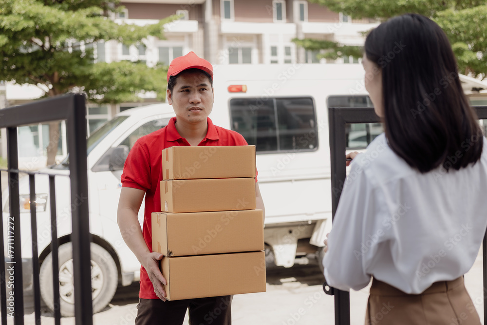 Fototapeta premium Young Asian woman receives a cardboard box from the postman at home who is smiling and happy. The courier delivers the item urgently. Online store, paper containers, postman, delivery service, parcels