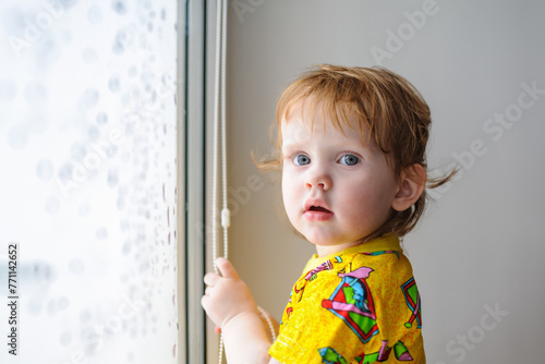 Little girl opening blinds of window at home.
