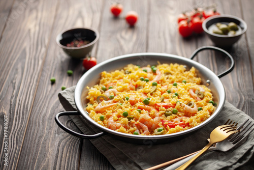 Traditional spanish seafood paella with rice, shrimps, olives and green peas in paellera on wooden background.