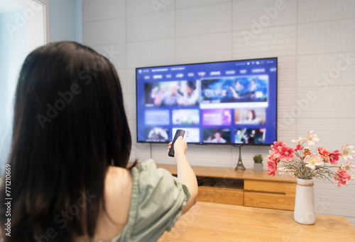An Asian woman is watching TV in the living room at home, holding a TV remote control
