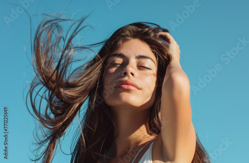 beautiful woman with long brown hair touching her shiny and smooth hair against a bright light blue background