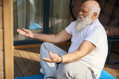 Old man sits in a yoga pose on a wooden veranda