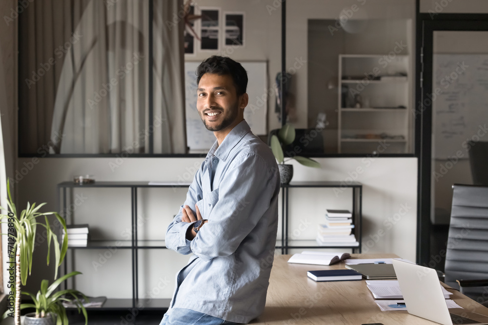 Happy confident handsome young Indian business man posing in office, standing at workplace table, looking at camera with hands crossed, smiling. Successful manager professional portrait