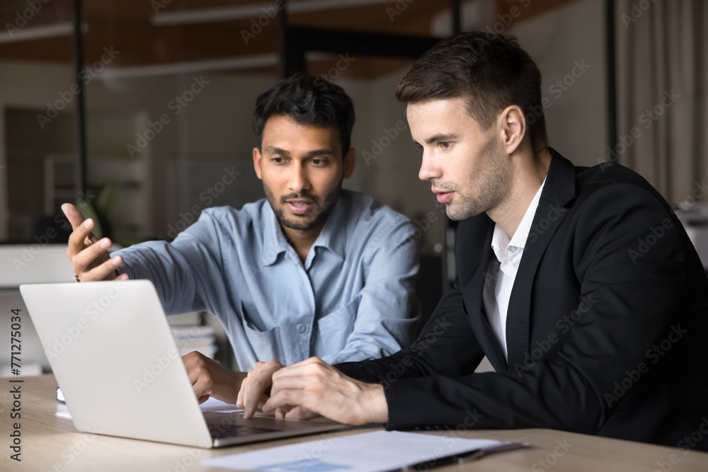 Two diverse business men talking at laptop, discussing online job application, Internet project, startup, looking, pointing at computer screen. Indian manager, presenting web content to customer Two diverse business men talking at laptop, discussing online job application, Internet project, startup, looking, pointing at computer screen. Indian manager, presenting web content to customer