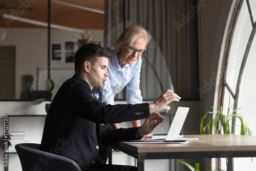 Two younger and elder business professionals discussing project tasks, working on startup plan, development strategy, watching content at laptop, moving hands at screen for explaining ideas