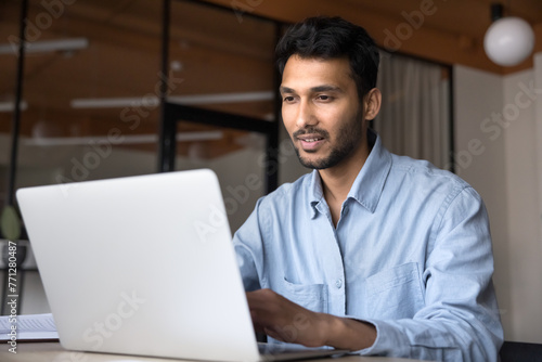 Positive young Indian business professional man in casual working at laptop in office, sitting at workplace table, typing, watching online video, using technology for job communication