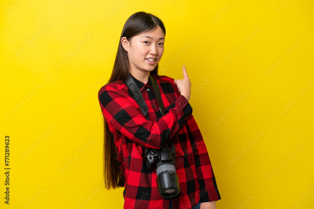 Young photographer Chinese woman isolated on yellow background pointing back Young photographer Chinese woman isolated on yellow background pointing back