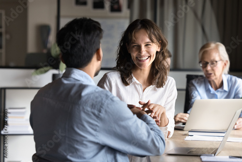 Cheerful young employee woman discussing project with colleague at meeting table, laughing, having fun, talking to coworkers. Two managers enjoying communication at office meeting