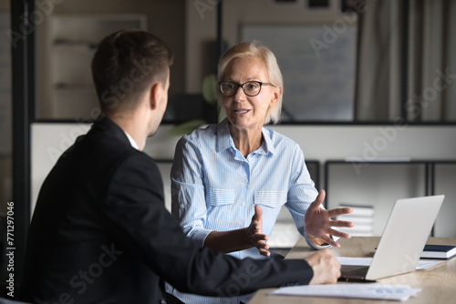 Foto Serious senior businesswoman and younger specialist man in formal suit talking at workplace, discussing job, project