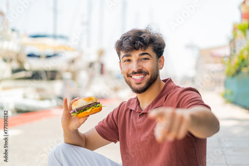Young Arabian handsome man holding a burger at outdoors points finger at you with a confident expression