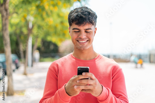Young hispanic man at outdoors sending a message or email with the mobile