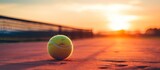 A vibrant tennis ball resting on the tennis court, bathed in the warm glow of the setting sun in the background