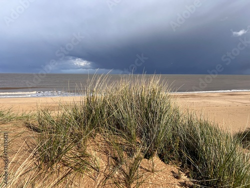 Man looking to ocean horizon beautiful sandy beach landscape grey stromy sky clouds and from grassy banks of sand dunes rolling waves on flat sea on Spring day in Winterton Norfolk East Anglia uk 