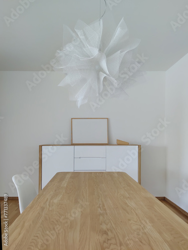 
Interior of a home. White tones and oak wood. Dining table, lamp and furniture with a blank photo frame to include a picture photo