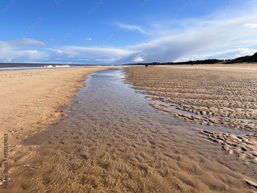 Stunning beautiful coast landscape of white sandy beach at low tide inlet river on shore to horizon with couple walking blue sky calm sea and white cloud Spring cold fresh day at Winterton Norfolk uk Stunning beautiful coast landscape of white sandy beach at low tide inlet river on shore to horizon with couple walking blue sky calm sea and white cloud Spring cold fresh day at Winterton Norfolk uk