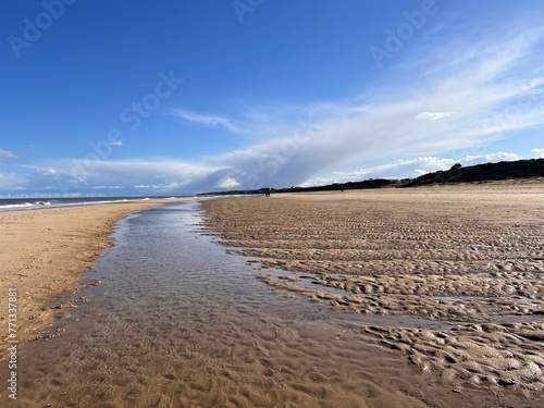 Stunning beautiful coast landscape of white sandy beach at low tide inlet river on shore to horizon with couple walking blue sky calm sea and white cloud Spring cold fresh day at Winterton Norfolk uk 