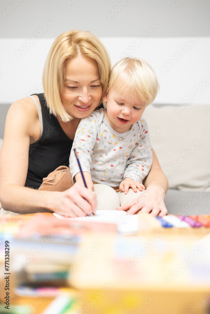 Caring young Caucasian mother and small son drawing painting in notebook at home together. Loving mom or nanny having fun learning and playing with her little 1,5 year old infant baby boy child Caring young Caucasian mother and small son drawing painting in notebook at home together. Loving mom or nanny having fun learning and playing with her little 1,5 year old infant baby boy child