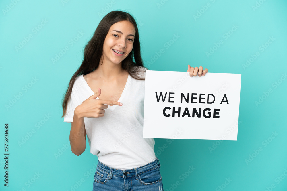 Young French woman isolated on blue background holding a placard with text We Need a Change and pointing it Young French woman isolated on blue background holding a placard with text We Need a Change and pointing it