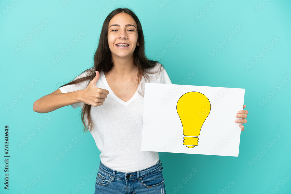 Young French woman isolated on blue background holding a placard with bulb icon with thumb up Young French woman isolated on blue background holding a placard with bulb icon with thumb up