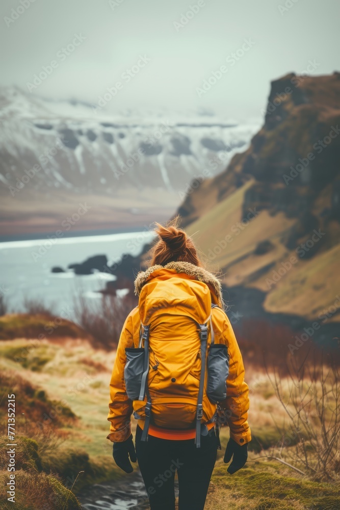 A woman in a yellow jacket with big backpack overlooking a scenic landscape of mountains, water, and grass.