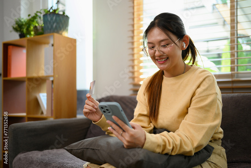 Pleased young Asian woman with credit card shopping online on mobile phone at home