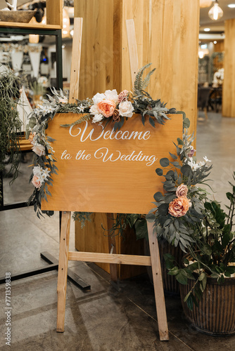 A wooden welcome sign covered in colorful flowers and lush greenery, creating a vibrant and inviting display. With the inscription "Welcome to the wedding"