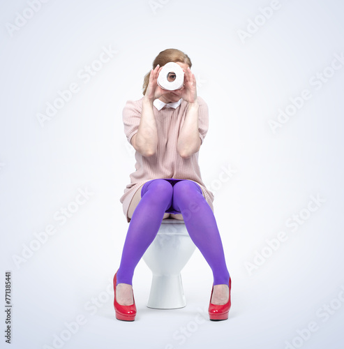 A young woman is sitting on the toilet and looking through a roll of toilet paper, isolated on a light blue background