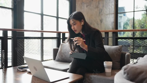 The young Asian businesswoman is taking notes on her tablet, participating in a video call business meeting via her laptop at the coffee shop photo