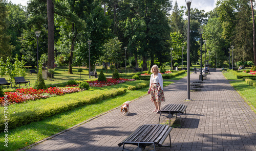 Woman and dog - Cavalier King Charles Spaniel - walking in a public park with lush flowers, plants, grass and trees